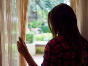 Rear view of a young woman holding the curtains open to look out of a large window in her apartment